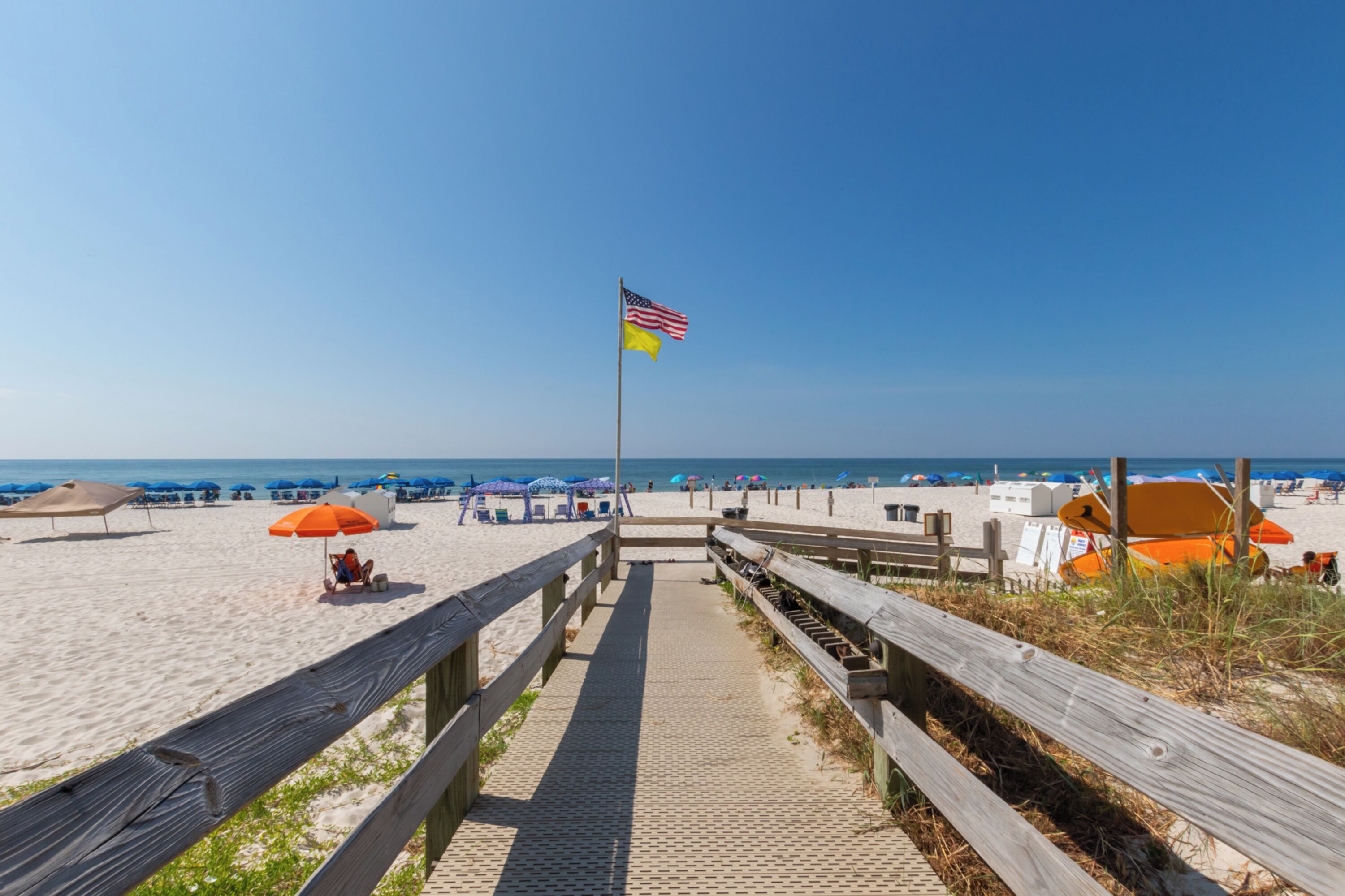 Ein Holzsteg führt zu einem Strand mit Sonnenschirmen und Liegestühlen.