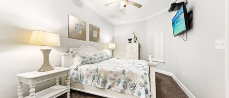 A bedroom with a bed, two bedside tables, a ceiling fan, a mounted TV, and a white dresser.