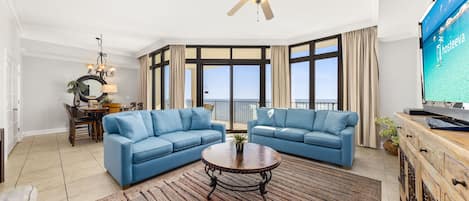 A spacious living room with two blue sofas, a wooden coffee table, a ceiling fan, and a large TV screen displaying 'Dhosteeva'.