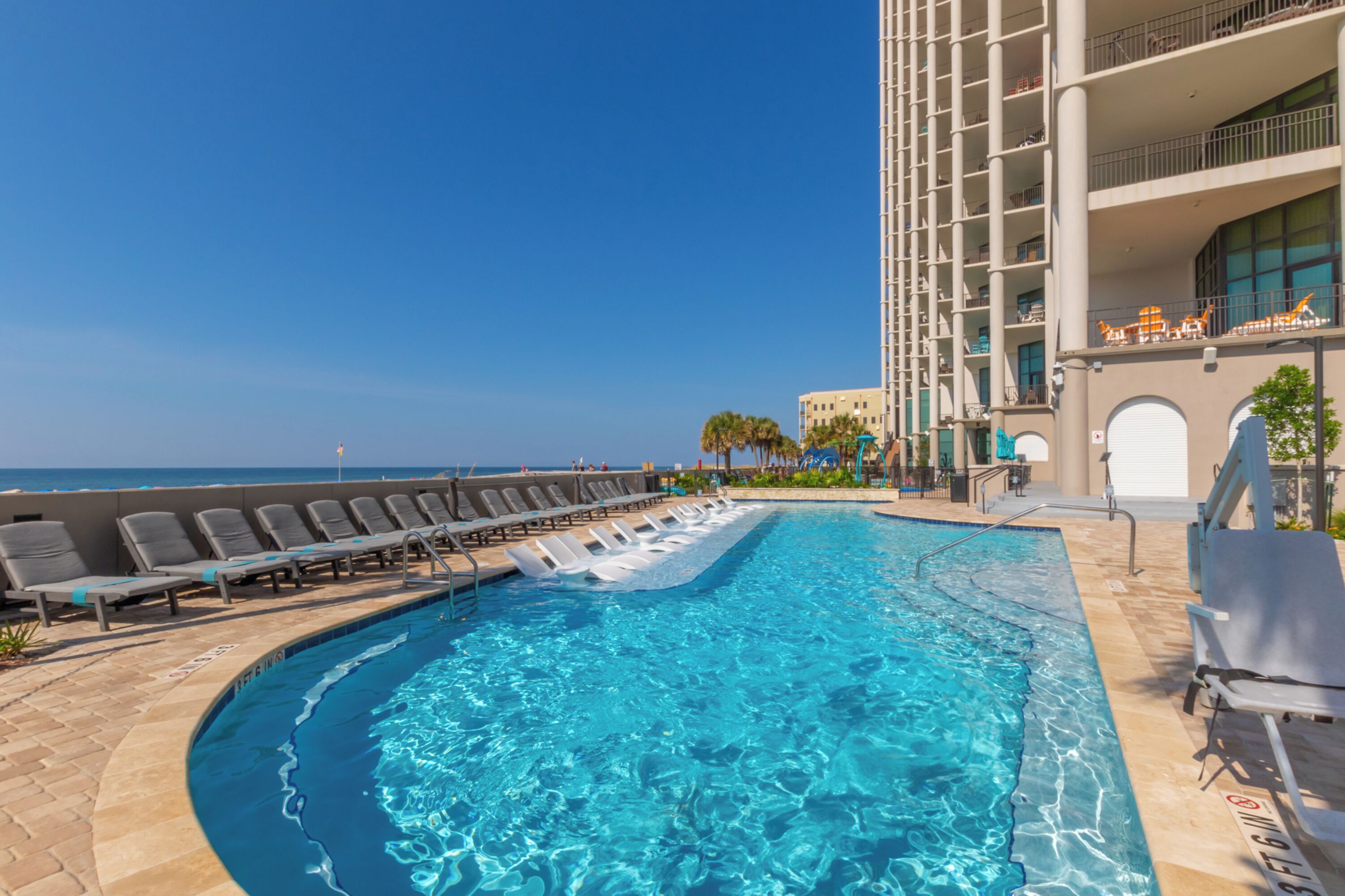 A swimming pool with lounge chairs, a building with balconies, and a clear sky.