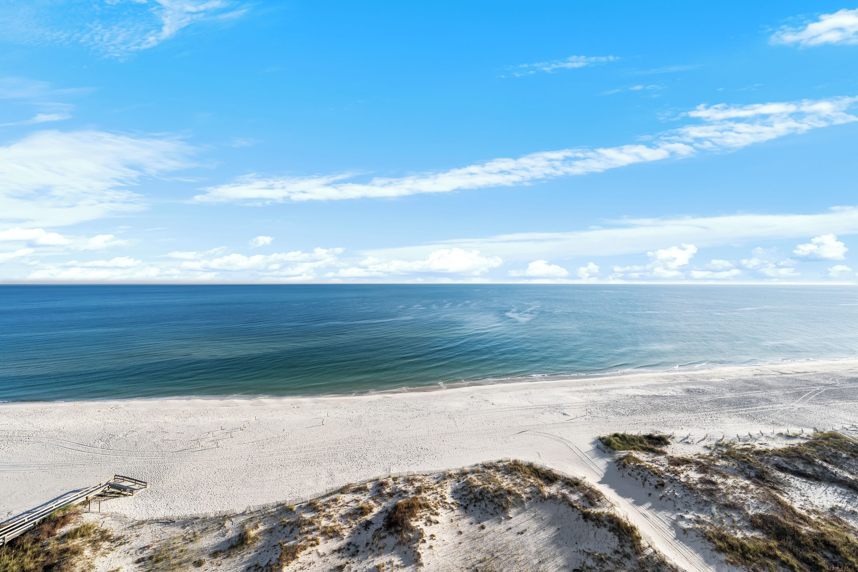 A beach with a wooden walkway leading to the water, sandy dunes, and a clear blue sky.