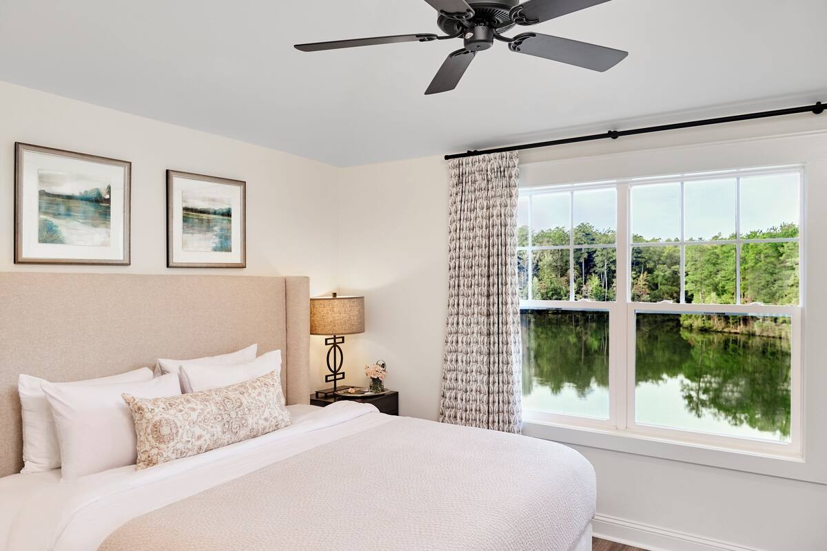 A bedroom with a bed, a ceiling fan, two framed pictures, a nightstand with a lamp, and a window with a view of greenery.