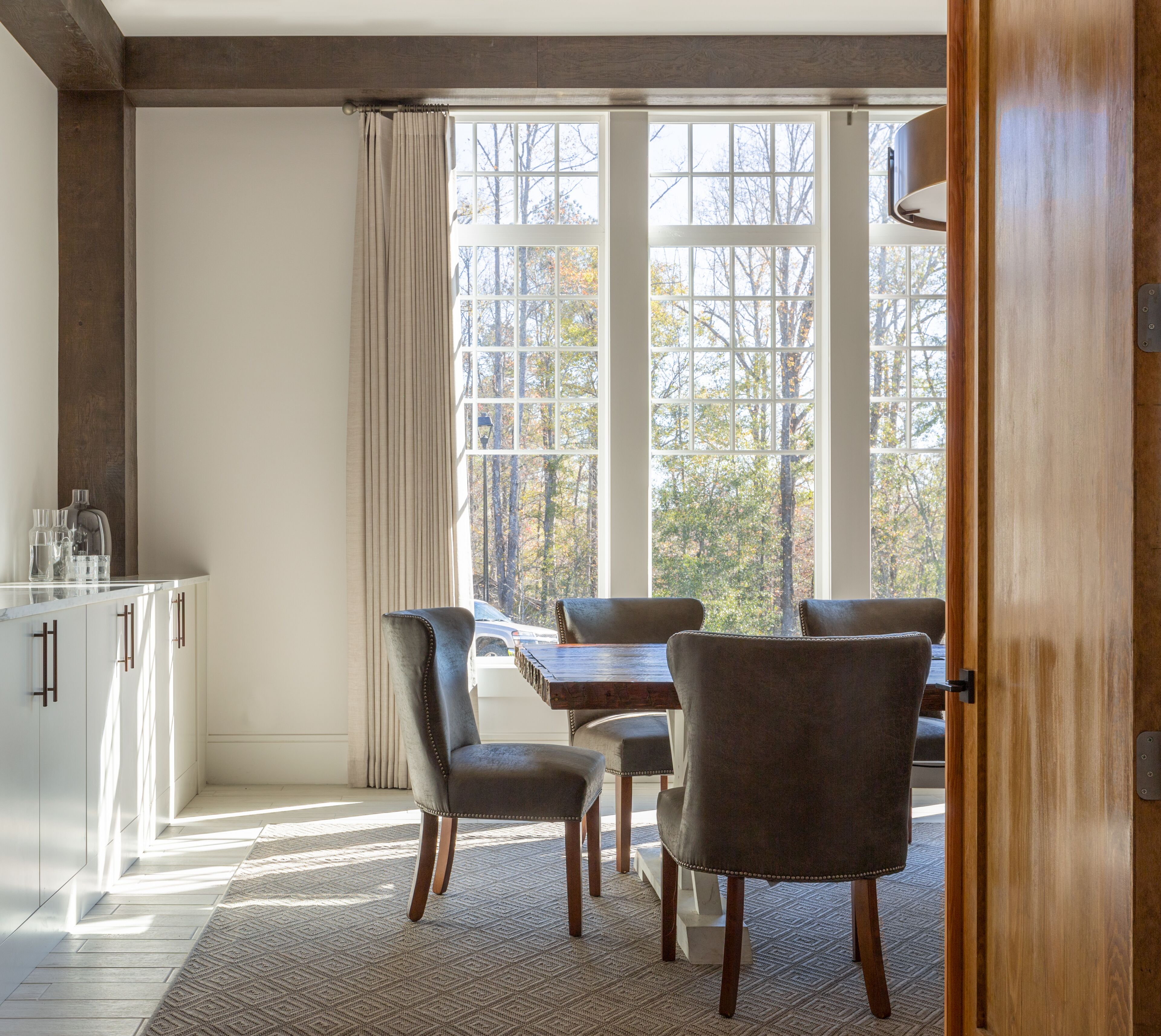 A dining area with a table and chairs, a kitchen with cabinets, and a view of trees through large windows.