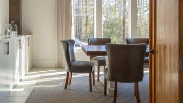 A dining area with a table and chairs, a kitchen with cabinets, and a view of trees through large windows.