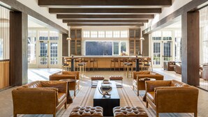 A spacious indoor area with leather sofas and ottomans arranged around a central table, large windows offering views of the outdoors, and a bar area with stools.