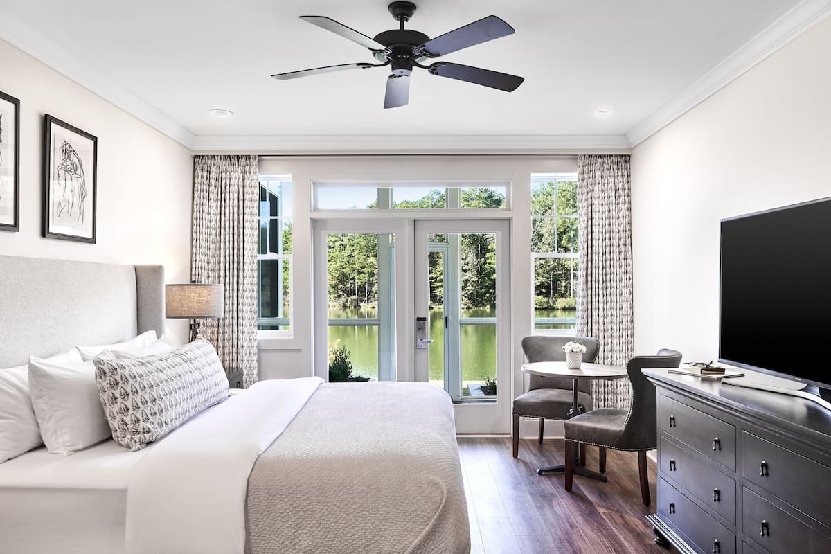 A bedroom with a bed, a desk, a chair, a TV, and a view of a lake through the window.