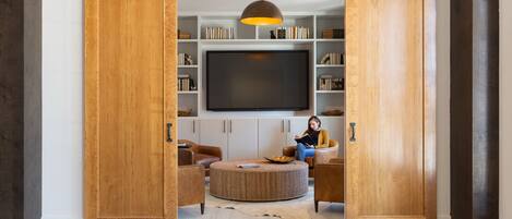 Una habitación con un gran armario corredero de madera, un televisor de pantalla plana, un otoman tejido redondo, sillones de cuero y una estantería con libros y adornos.