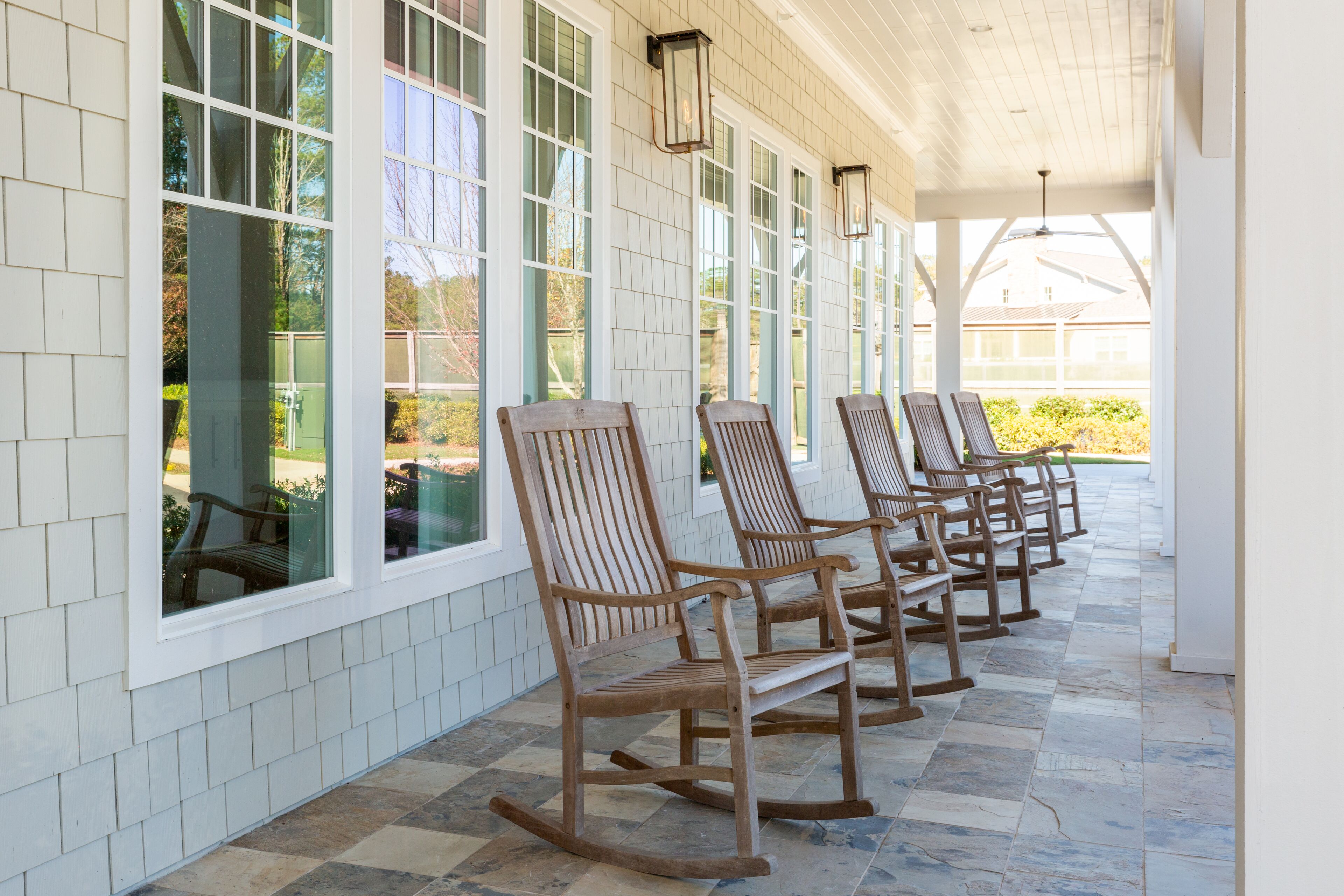 A covered porch with wooden rocking chairs aligned against a white exterior wall with large windows.