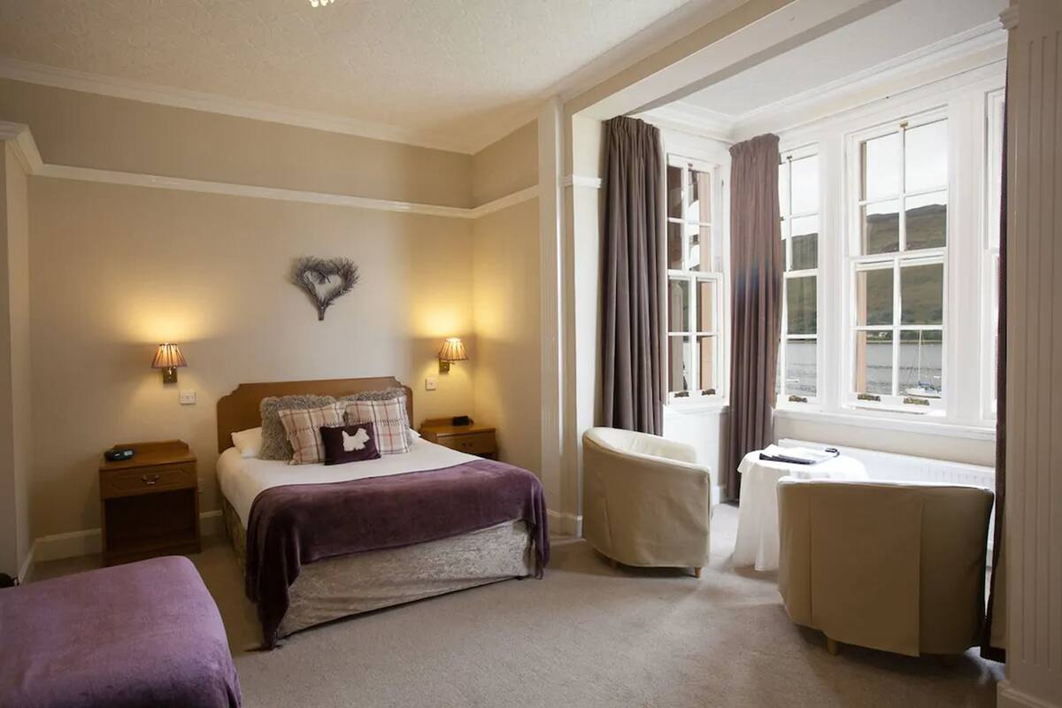 Habitación de hotel con cama, mesitas de noche, escritorio y una silla. Hay cortinas en las ventanas y una decoración con forma de corazón en la pared.