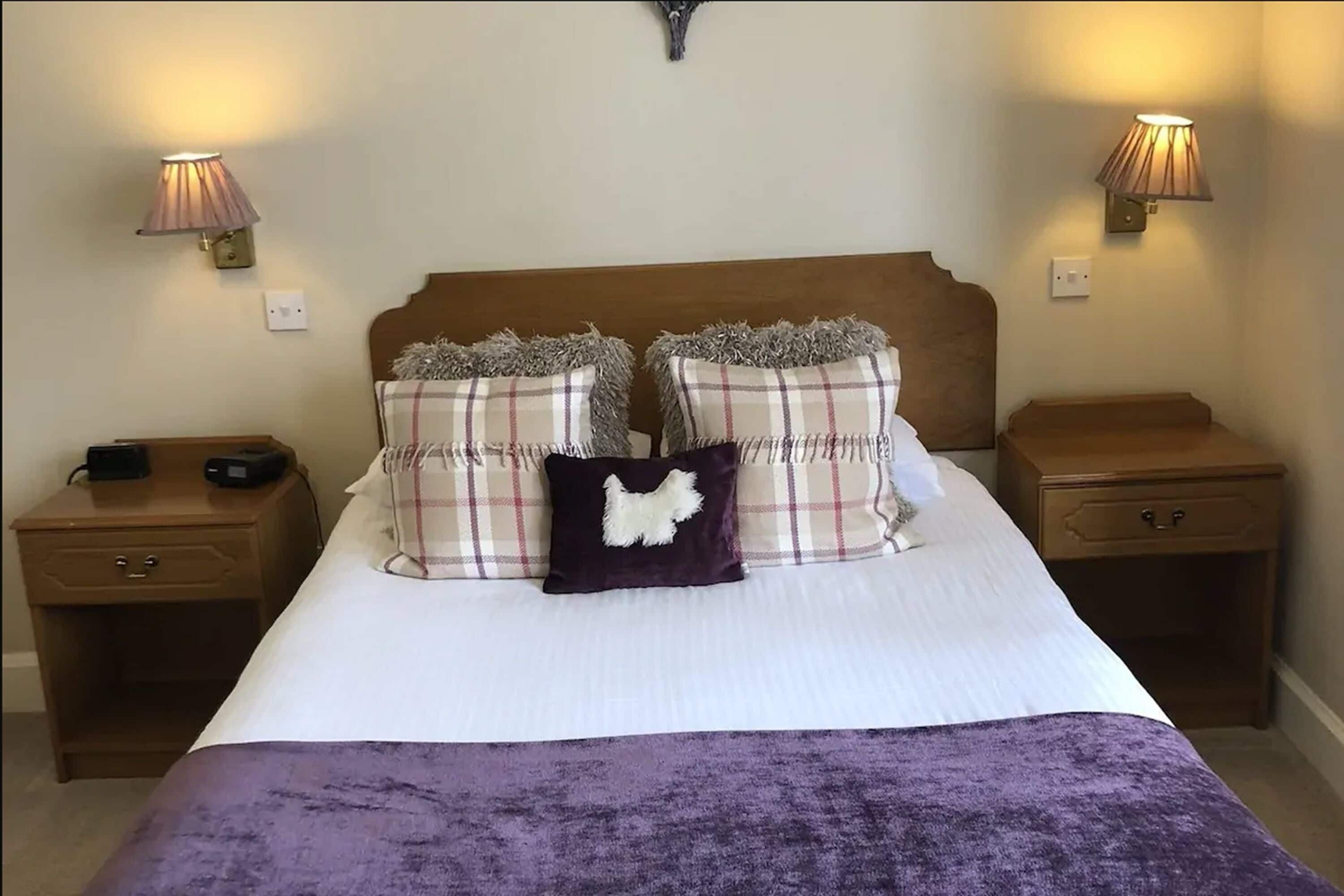 Una cama bien hecha con edredón blanco, dos almohadas escocesas y una manta color morado. Hay dos mesitas de noche con lámparas, y un pequeño aparador a la izquierda.