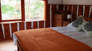 A bedroom with a wooden headboard, a large window, a bed with an orange bedspread, and a nightstand with a lamp. - Lodge Palguin Alto (Pucón)