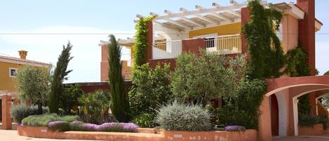 A multi-story building with a covered patio area, surrounded by landscaped gardens and various plants.