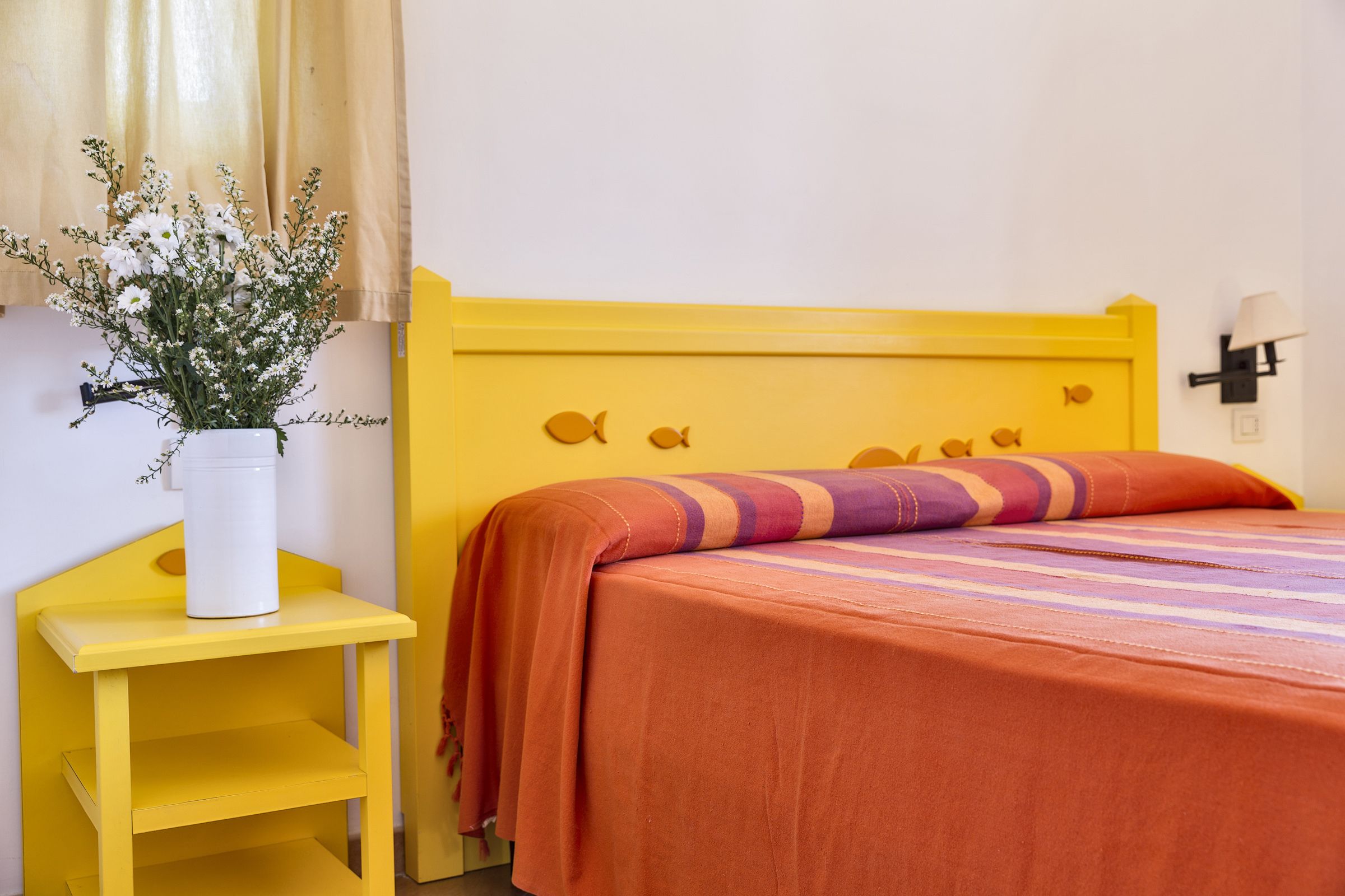 A yellow bed with a striped blanket and a white vase with flowers on a yellow nightstand.