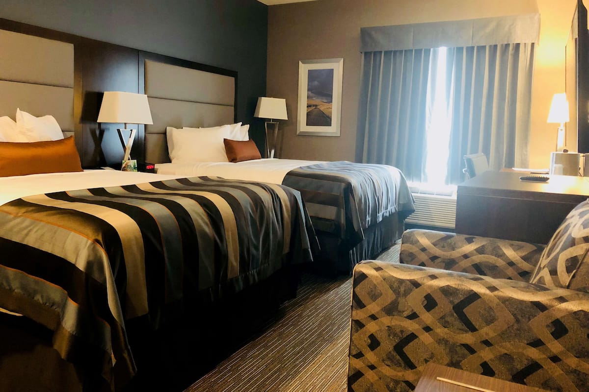 Habitación de hotel con dos camas, un escritorio, una silla y un gran ventanal con cortinas.
