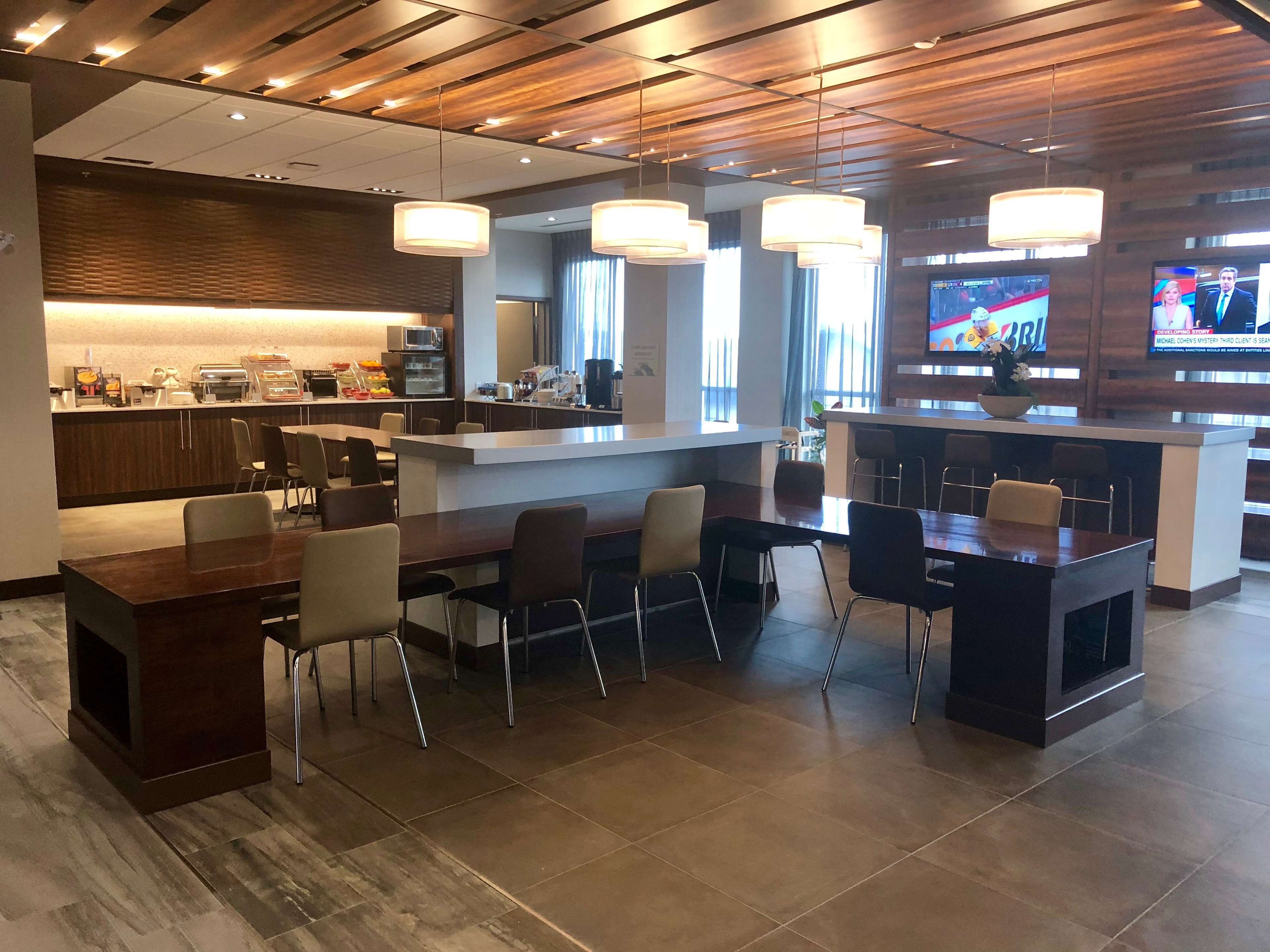 A modern hotel lobby with a kitchen area, dining tables, and a TV screen displaying a program.