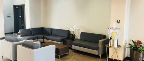 A modern waiting area with a sectional sofa, a white armchair, and a small table. A water dispenser is on a stand near the wall.