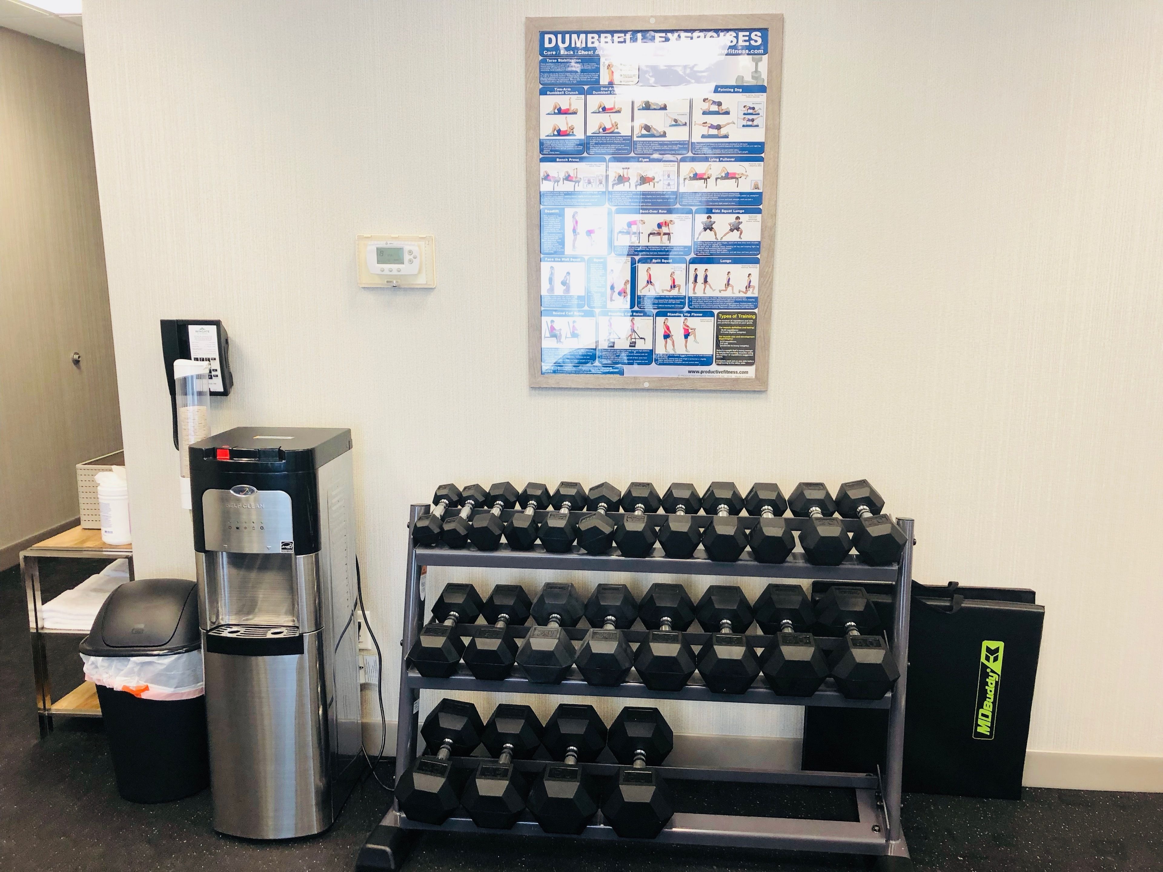 A gym area with a water dispenser, a coffee machine, a trash bin, and a rack of dumbbells.