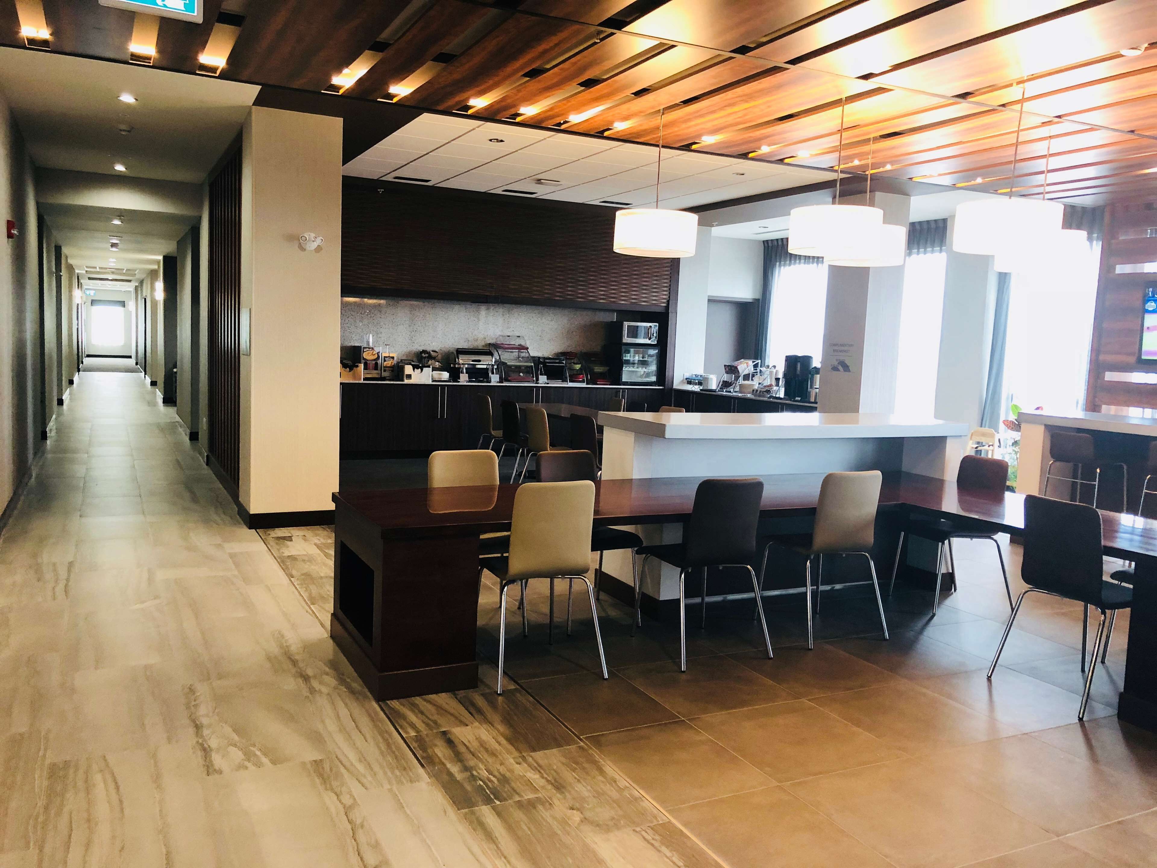 A modern hotel lobby with a long corridor, a central bar area with seating, and a kitchen in the background.