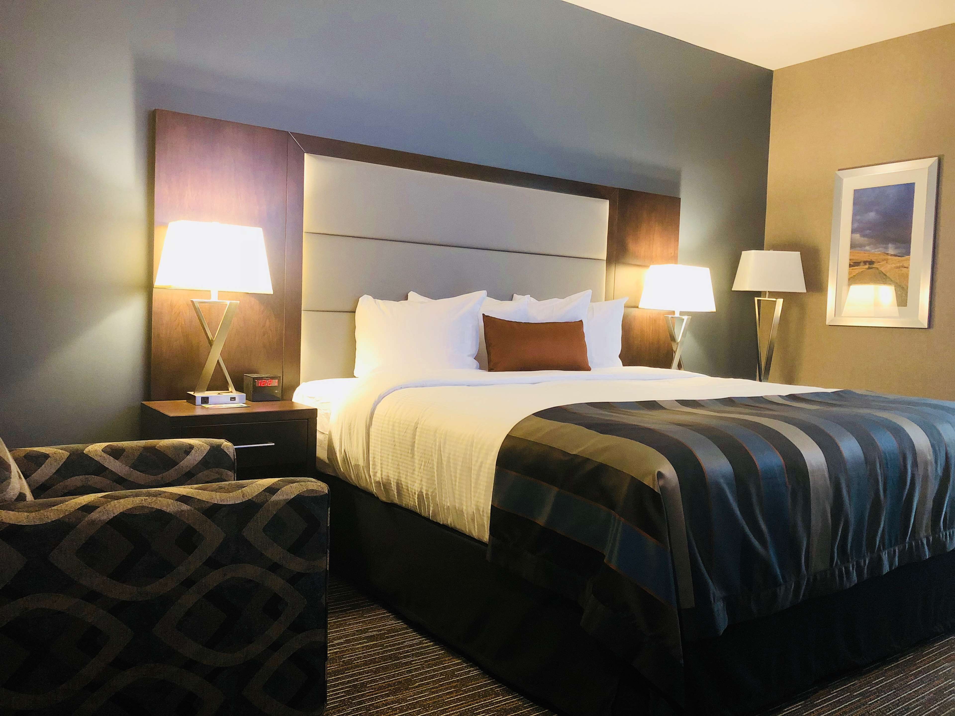 A hotel room with a large bed, two bedside tables with lamps, a patterned armchair, and a framed picture on the wall.