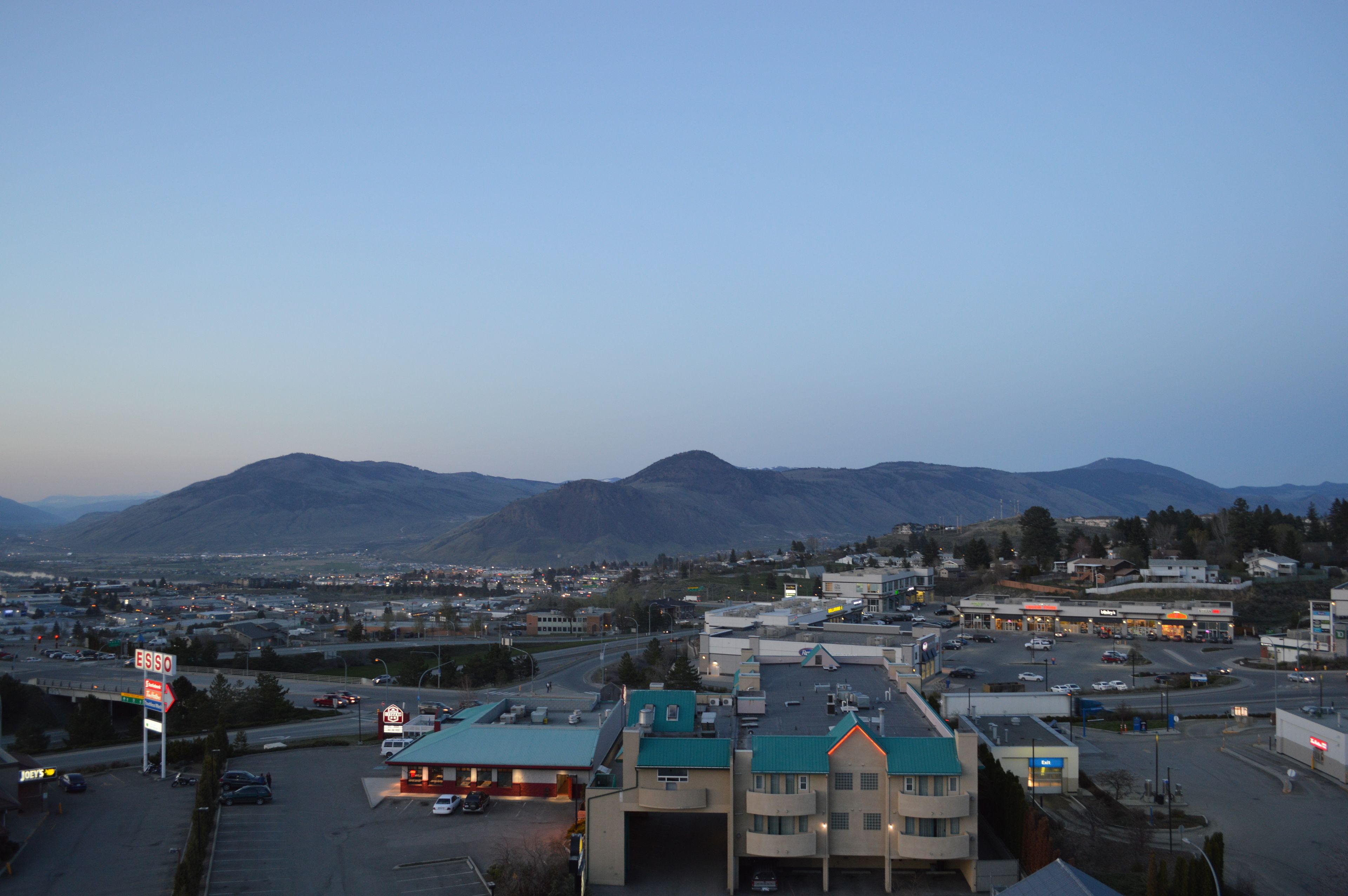 A town with a gas station, a large building with a green roof, and mountains in the background.