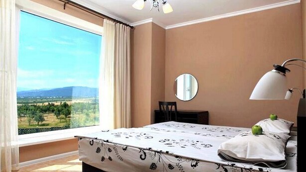 Un dormitorio con una ventana grande, una cama, una mesita de noche, una lámpara y un espejo.