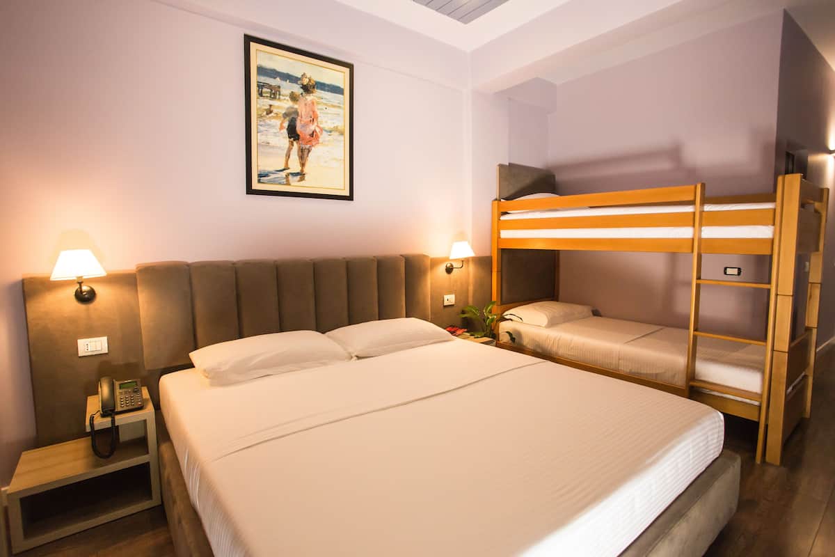 A hotel room with a large bed, a bunk bed, bedside lamps, a painting on the wall, and a nightstand with a phone.