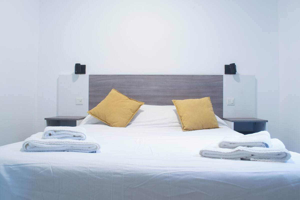 A neatly made bed with white linens and two yellow pillows, flanked by two bedside tables with black lamps.