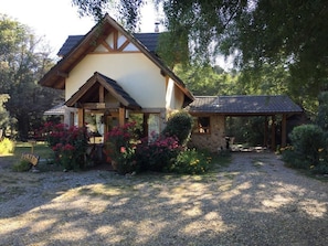 Uma casa de madeira tradicional com uma extensão em pedra, cercada por um caminho de cascalho e vegetação exuberante.
