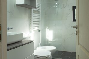 A modern bathroom with a white sink, toilet, and bidet, marble walls, and a glass shower enclosure. - Cabanas Duendes del Maiten (San Carlos de Bariloche)