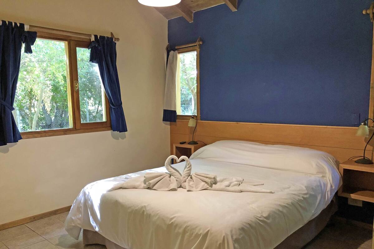 A hotel room with a bed, two windows with curtains, a nightstand with a lamp, and a wooden headboard.