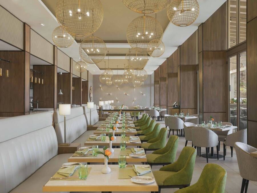 Un amplio comedor con varias mesas, sillas tapizadas de verde y lámparas esféricas colgantes.