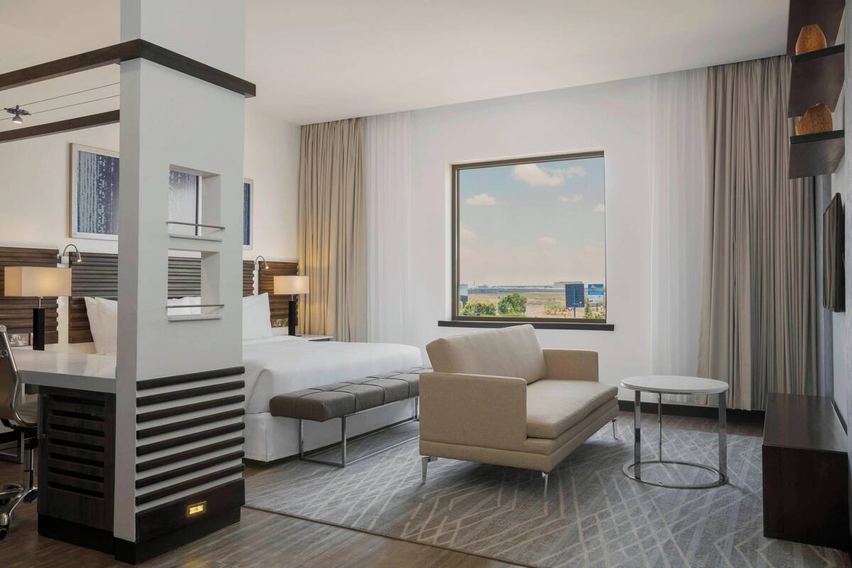Habitación de hotel con cama, sofá, una mesita y un escritorio con silla. Amplia ventana con vista a la ciudad.