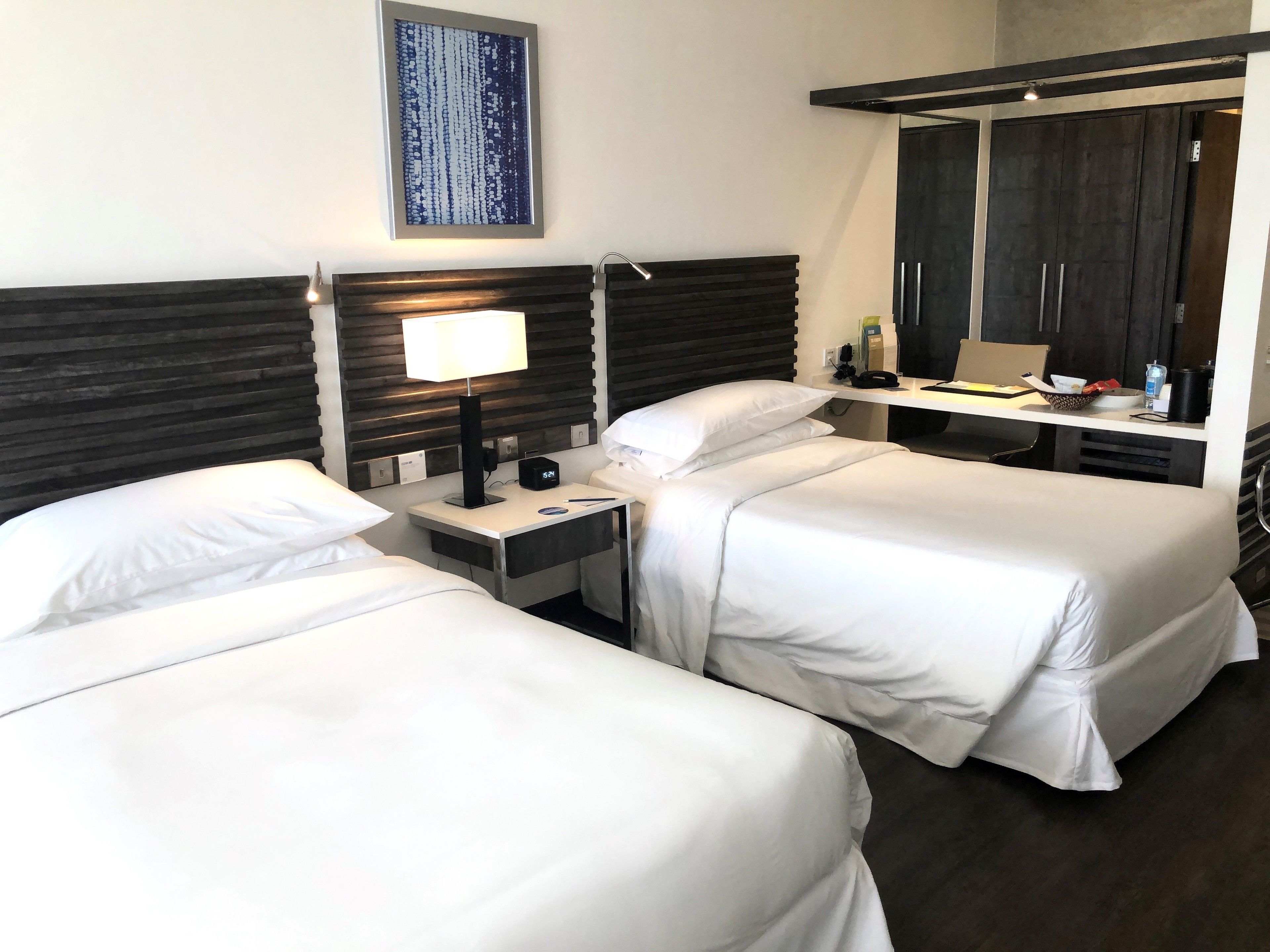 Une chambre d’hôtel avec deux lits simples, une table de chevet avec une lampe, un petit bureau avec un téléphone et un bol, et un grand miroir au-dessus du bureau.