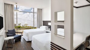 A hotel room with two beds, a small table, a chair, a desk with a lamp, and a large window offering a view of the outdoors.