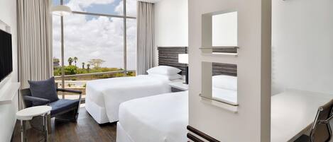 A hotel room with two beds, a small table, a chair, a desk with a lamp, and a large window offering a view of the outdoors.