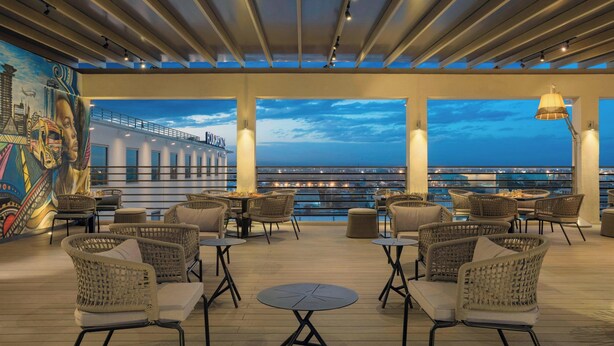 A rooftop lounge with wicker furniture, a mural, and a view of the cityscape at dusk.