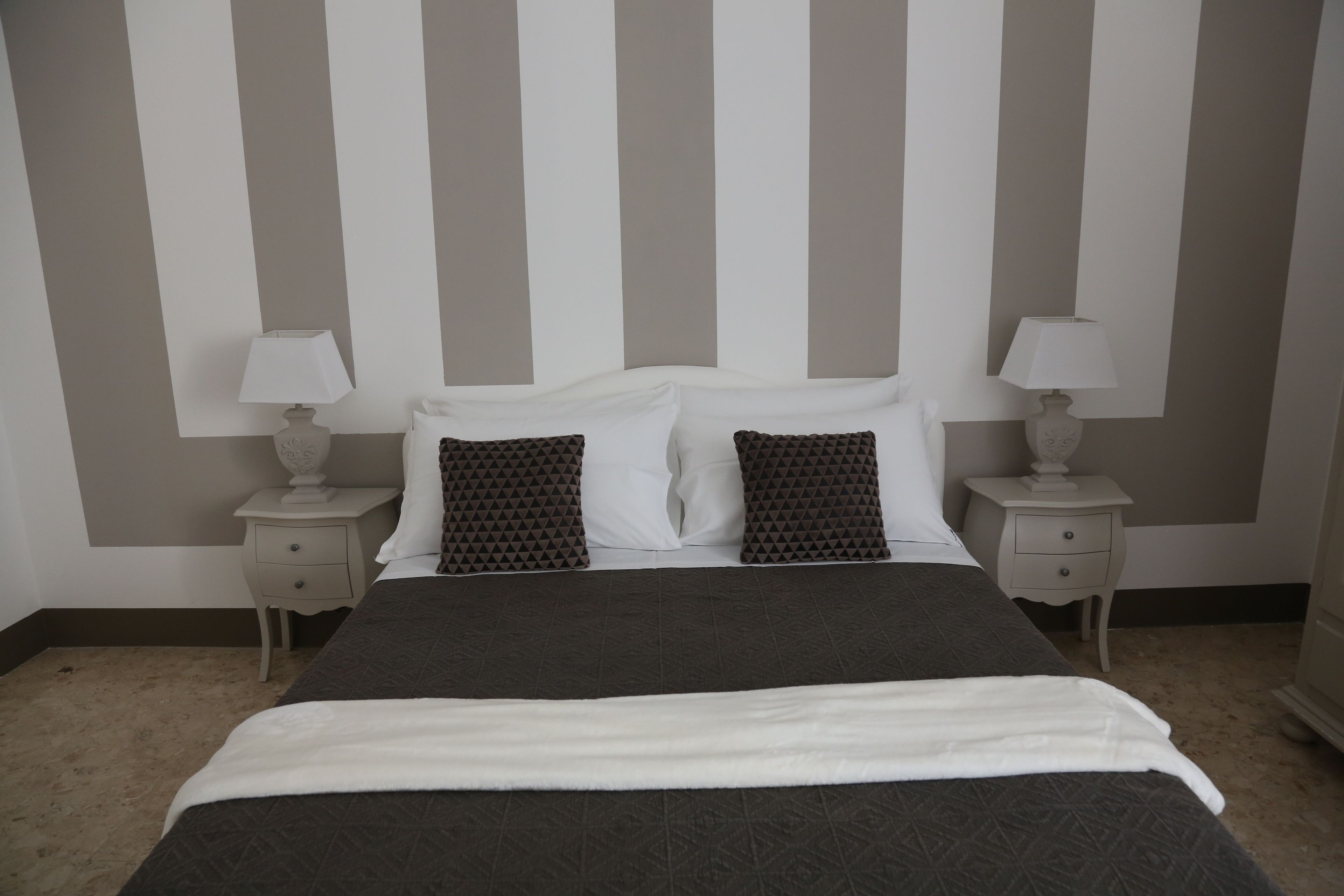 A neatly made bed with a dark patterned bedspread and white pillows, flanked by two bedside tables with lamps, in a room with striped walls.