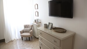 A room with a beige armchair, a white dresser with drawers, a flat-screen TV mounted on the wall, and a small table with a coffee pot and picture frames. - Dimi House (Lecce)