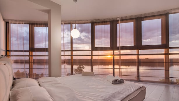 Une chambre avec un grand lit, un luminaire suspendu et une vue sur un coucher de soleil se reflétant sur une étendue d’eau, visible à travers de grandes fenêtres.