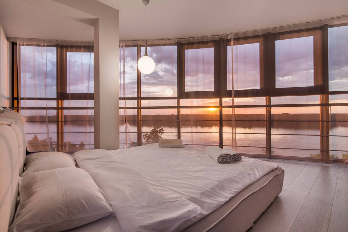 A bedroom with a large bed, a hanging light fixture, and a view of a sunset over a body of water through large windows.