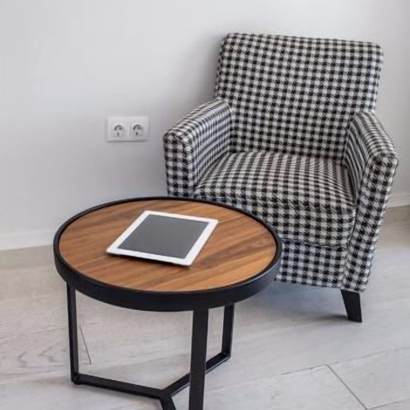 A round wooden table with a tablet on it, positioned in front of a houndstooth patterned armchair.