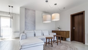 Un salon moderne avec un canapé d’angle, deux tabourets en bois, un tapis rond et des luminaires suspendus.