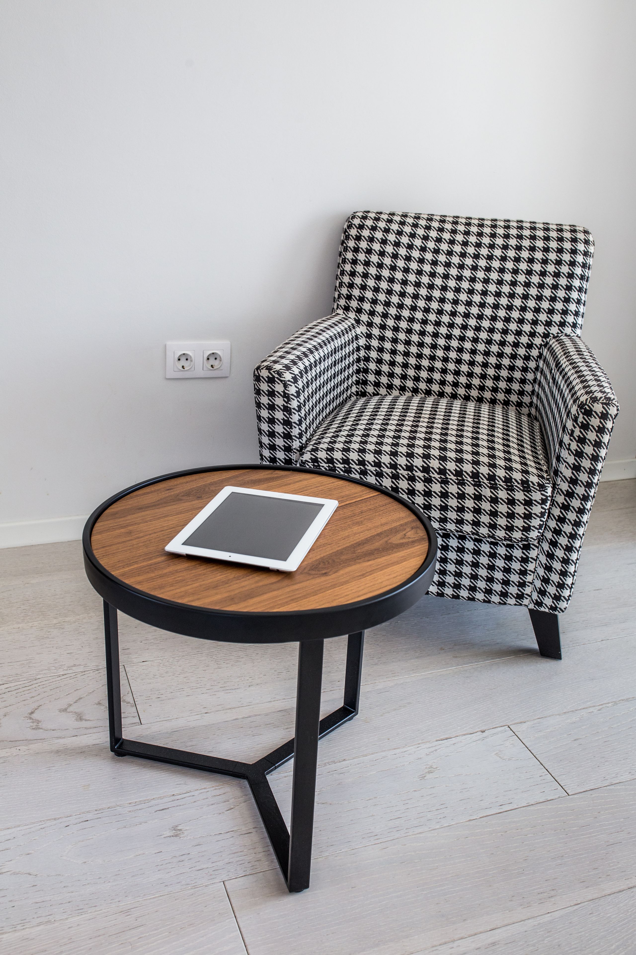 A round wooden table with a tablet on it, positioned next to a houndstooth patterned armchair in a room with a light-colored floor and a white wall.
