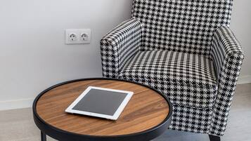 A round wooden table with a tablet on it, positioned next to a houndstooth patterned armchair in a room with a light-colored floor and a white wall.