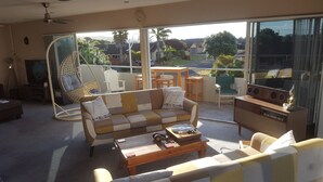Una sala de estar con sofá, mesa de centro y televisor. Hay un balcón con vistas a casas y árboles.