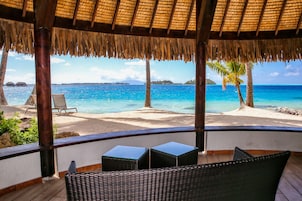 A beach view from a covered area with wicker furniture and a thatched roof.