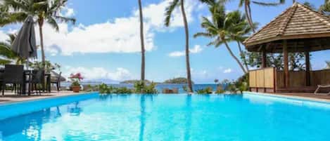 A clear blue pool surrounded by palm trees and a thatched-roof gazebo.