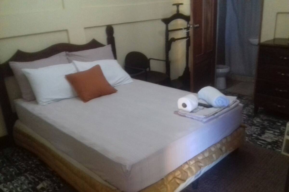 A bed with a wooden headboard, a brown pillow, and a white pillow. There is a small table with a towel and a bottle on it.