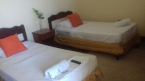 Dos camas individuales con sábanas blancas y almohadas naranjas, una mesita de noche con un florero, y piso alfombrado.