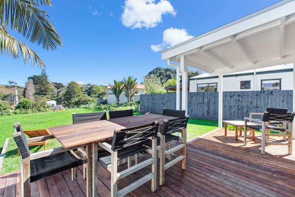 An outdoor patio with a wooden deck, a covered seating area, and a view of the surrounding greenery and houses. - The Little Blue Bach in Oneroa Village (Waiheke Island)